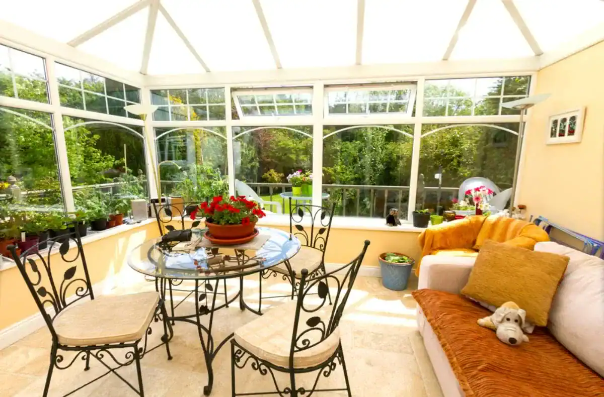 A bright sunroom in NY with large glass windows, a round glass table with four chairs, potted plants, a cozy sofa with cushions and a stuffed dog toy, overlooking a lush green garden&mdash;a perfect example of sunrooms Long Island style.